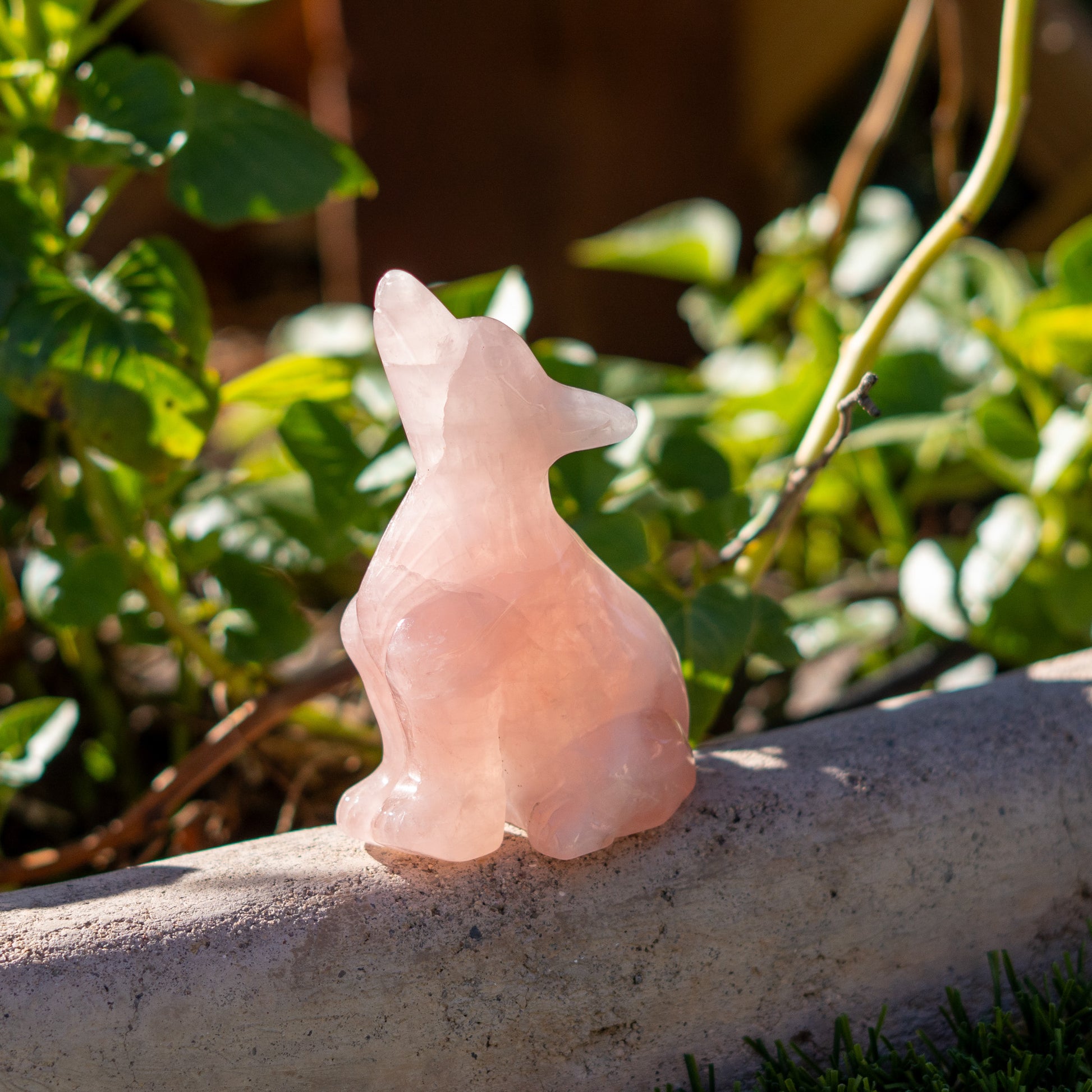 Pink stone sculpture of a cat on a stone ledge with green foliage in the background