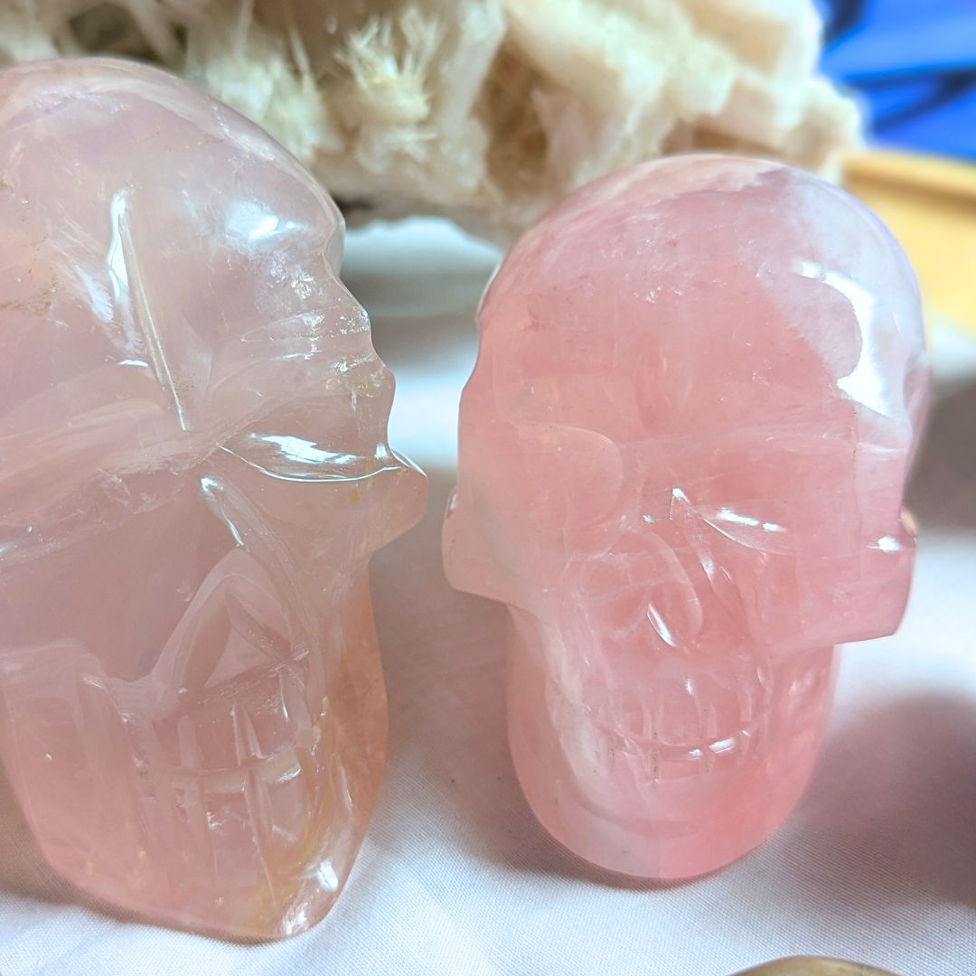 Two translucent pink rose quartz crystal skull carvings, one slightly larger, on a white fabric.